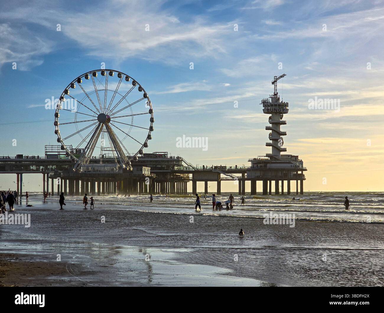 Les gens se promènent le long d'une plage de sable près d'une jetée avec une grande roue et une tour, baignés dans la douce lueur d'un coucher de soleil côtier coloré Banque D'Images