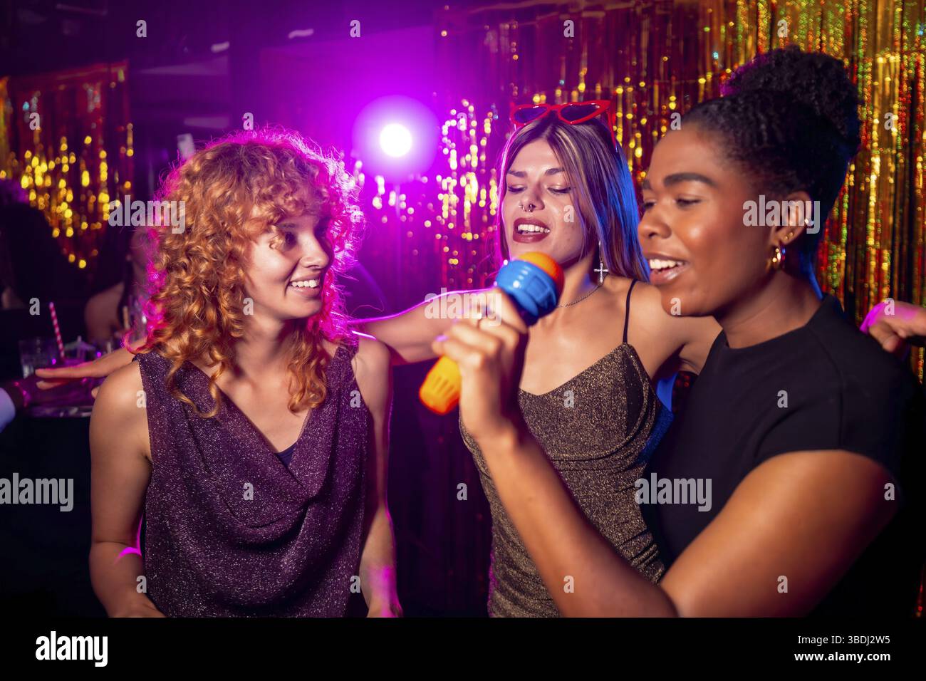 Amies féminines multi ethniques joyeuses chantant karaoké, profitant de la vie nocturne et s'amusant ensemble dans une boîte de nuit Banque D'Images