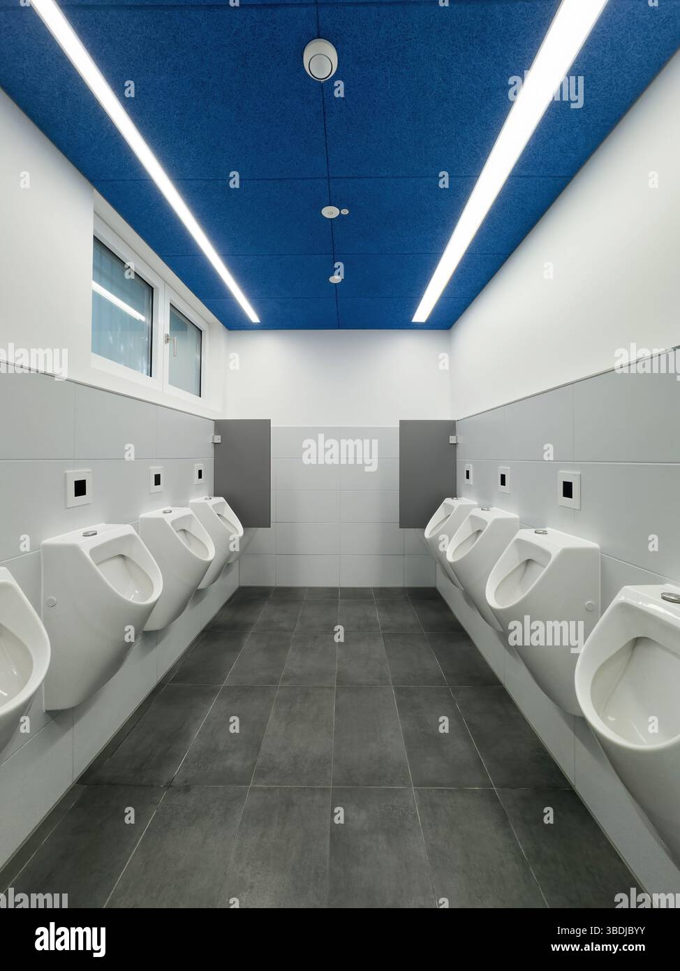 Berlin, Allemagne - 12 avril 2025 : vue symétrique d'un intérieur de toilettes moderne et épuré avec un plafond acoustique bleu, des urinoirs muraux, des surfaces carrelées et des lampadaires lumineux Banque D'Images Berlin, Allemagne - 12 avril 2025 : vue symétrique d'un intérieur de toilettes moderne et épuré avec un plafond acoustique bleu, des urinoirs muraux, des surfaces carrelées et des lampadaires lumineux Banque D'Images