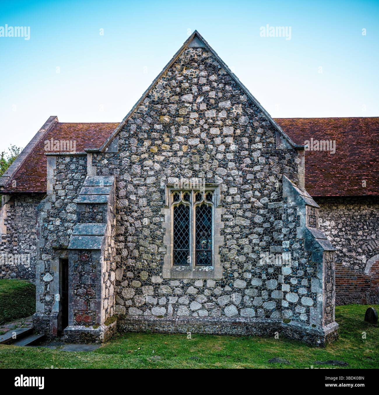 Détail d'un mur d'église en silex montrant une fenêtre gothique en verre plombé adjacente à une petite structure en pierre saillante avec un toit en tuiles, peut-être un vestr Banque D'Images