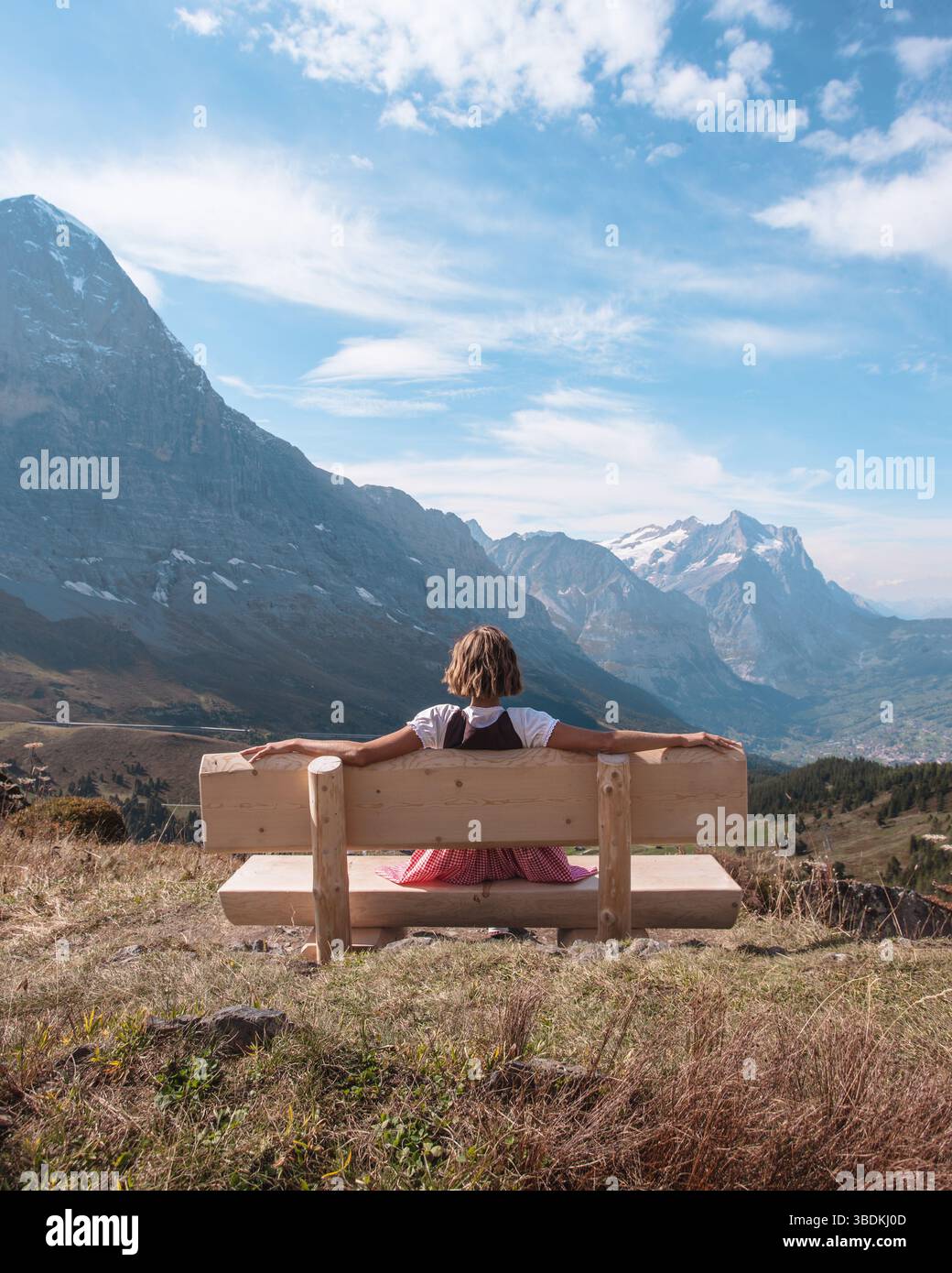 Belle jeune femme portant des vêtements traditionnels bavarois - dirndl ou tracht assis sur le banc dos à la caméra et regardant le paysage des Alpes européennes. Banque D'Images