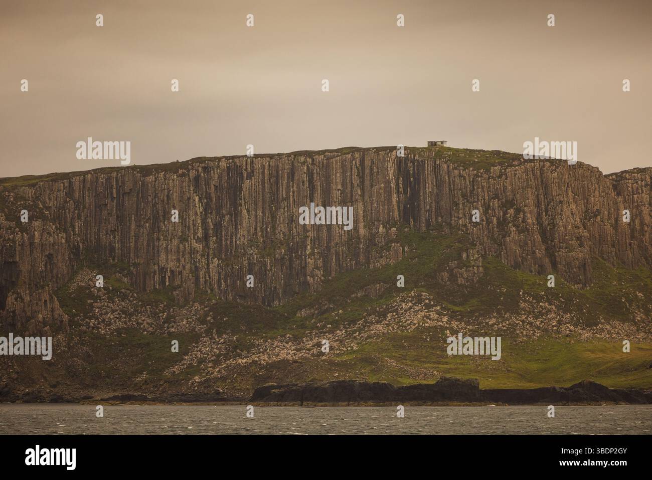 Les falaises verticales de basalte s'élèvent abruptement au-dessus des pentes rocheuses de l'île de Skye, en Écosse, avec un petit bâtiment visible sur le plateau sommital. Banque D'Images Les falaises verticales de basalte s'élèvent abruptement au-dessus des pentes rocheuses de l'île de Skye, en Écosse, avec un petit bâtiment visible sur le plateau sommital. Banque D'Images