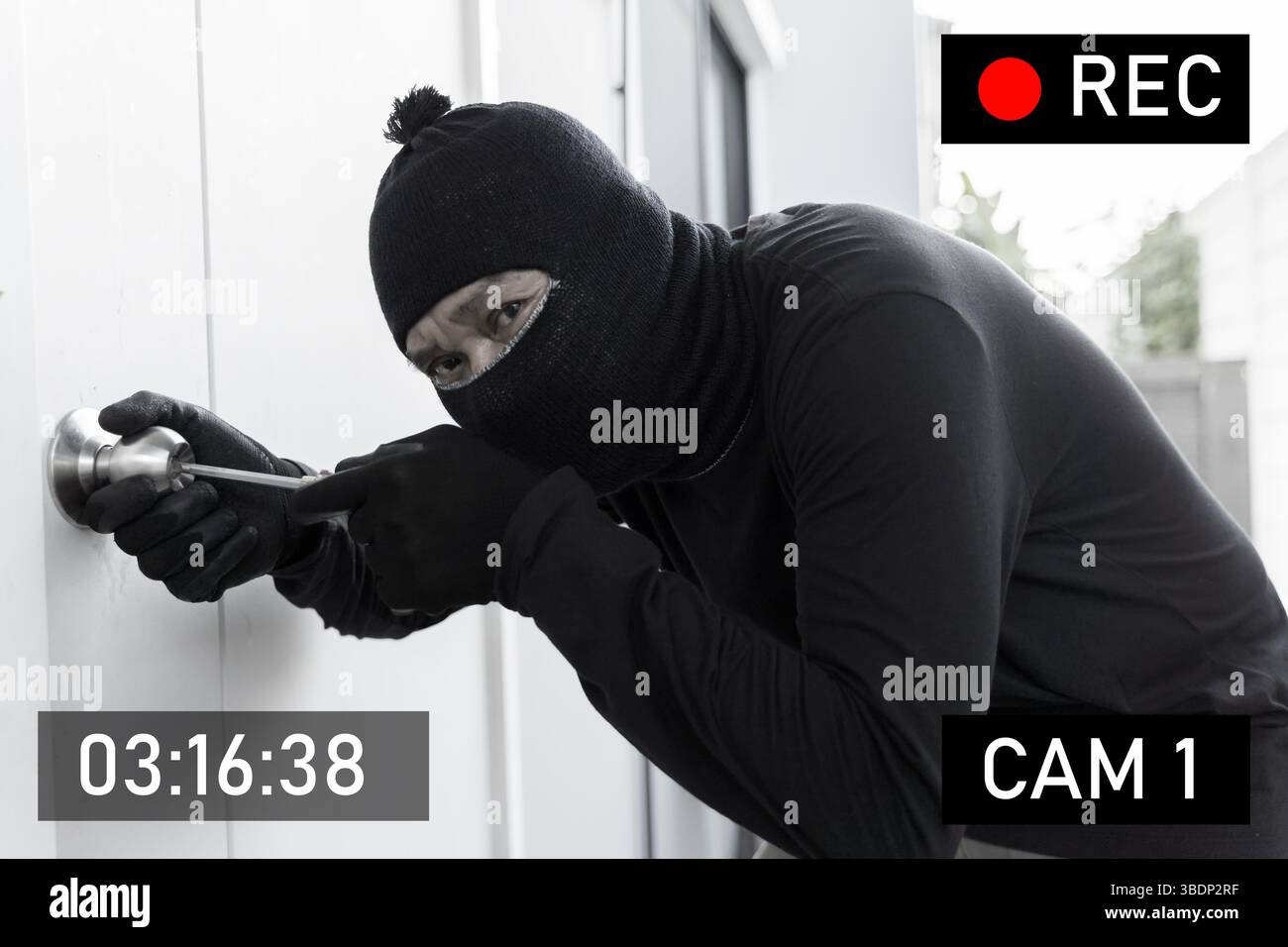 Un voleur masqué en gants noirs est vu à l'aide d'un tournevis pour pénétrer dans une porte de maison, capturé par la caméra de sécurité. Concept de cambriolage, invasion de domicile Banque D'Images