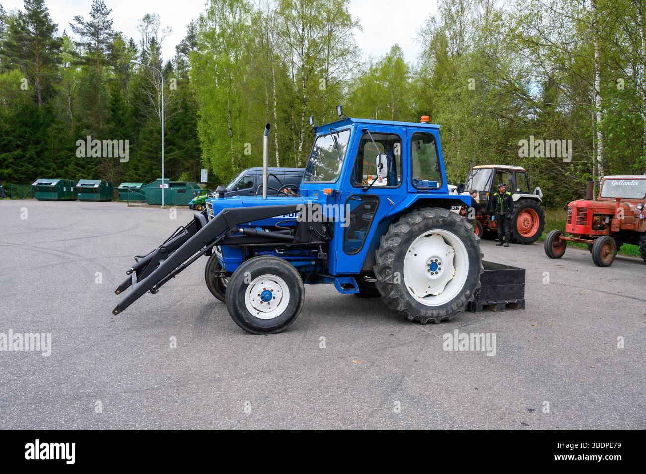 Un tracteur bleu vif est stationné dans un terrain de gravier, avec des arbres verts en arrière-plan et divers autres tracteurs à proximité. C'est un jour clair, mettant en valeur t Banque D'Images
