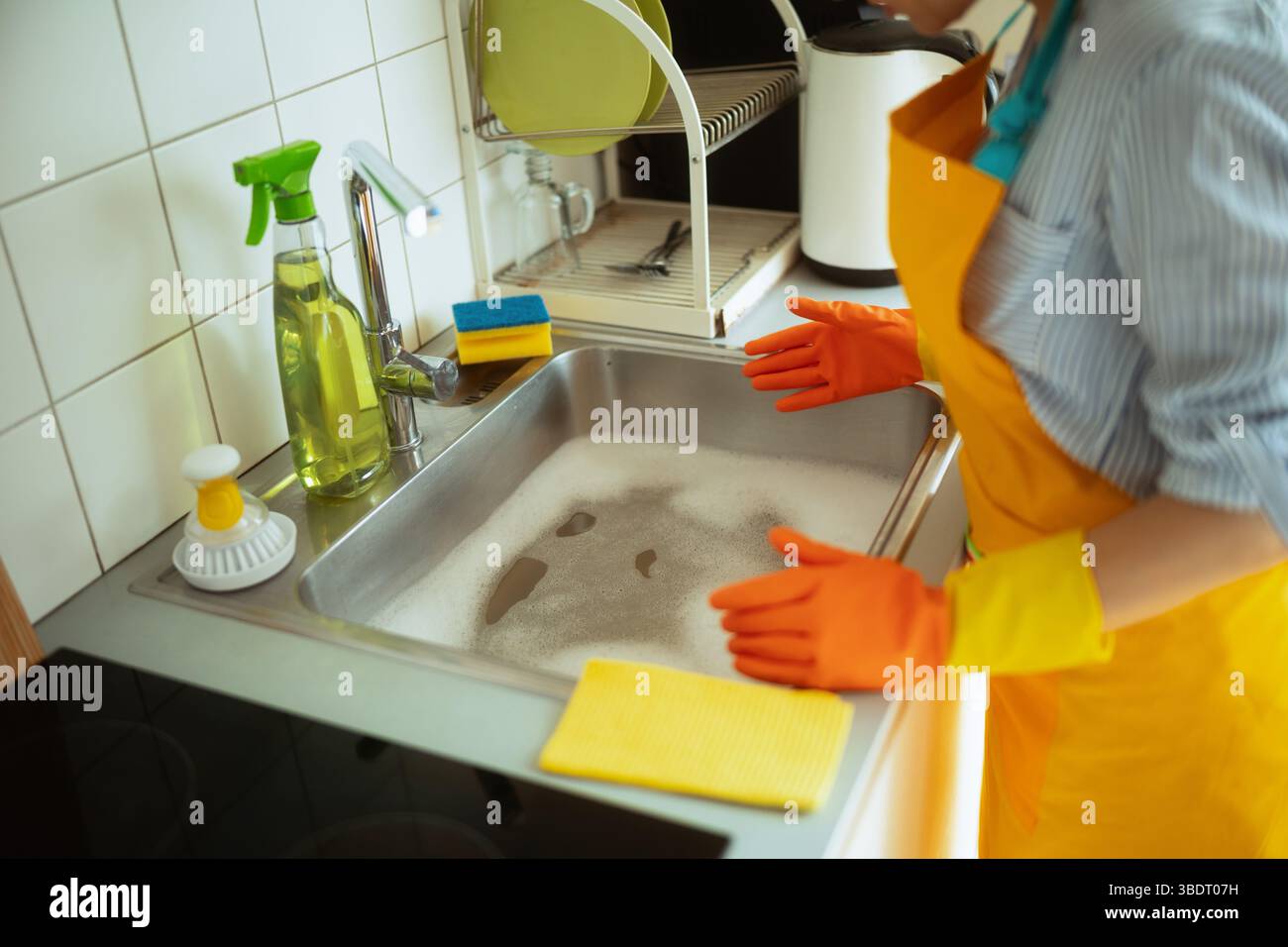 Une femme à un évier de cuisine rempli d'eau savonneuse, les mains levées, faisant face à un problème possible de plomberie ou de drainage tout en étant entourée de produits de nettoyage. Banque D'Images