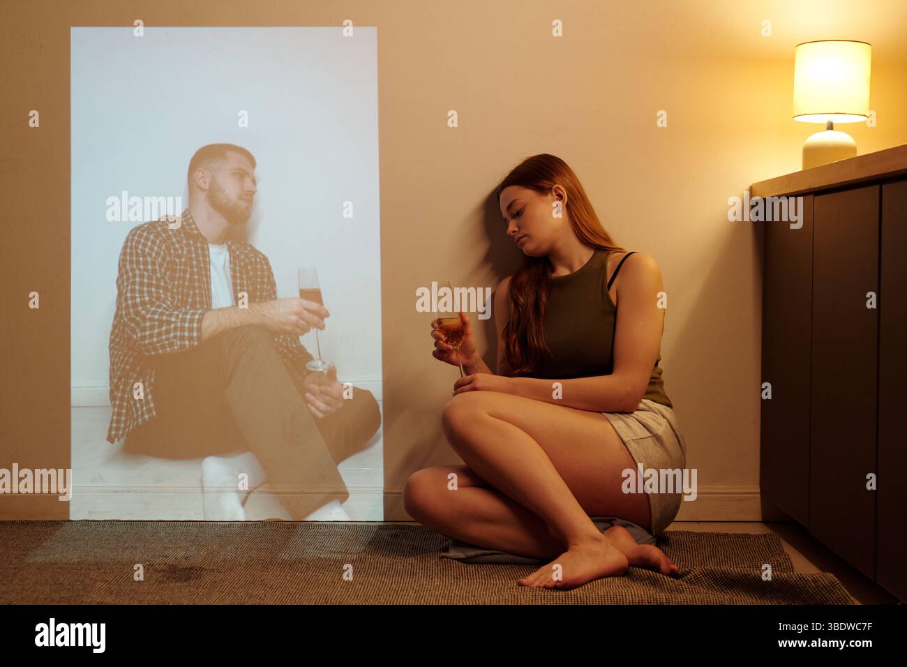 Femme assise sur le sol avec boisson, à côté de la lampe allumée, projetant l'image de l'homme buvant sur le mur. Le cadre semble confortable avec un éclairage chaleureux et une atmosphère détendue Banque D'Images
