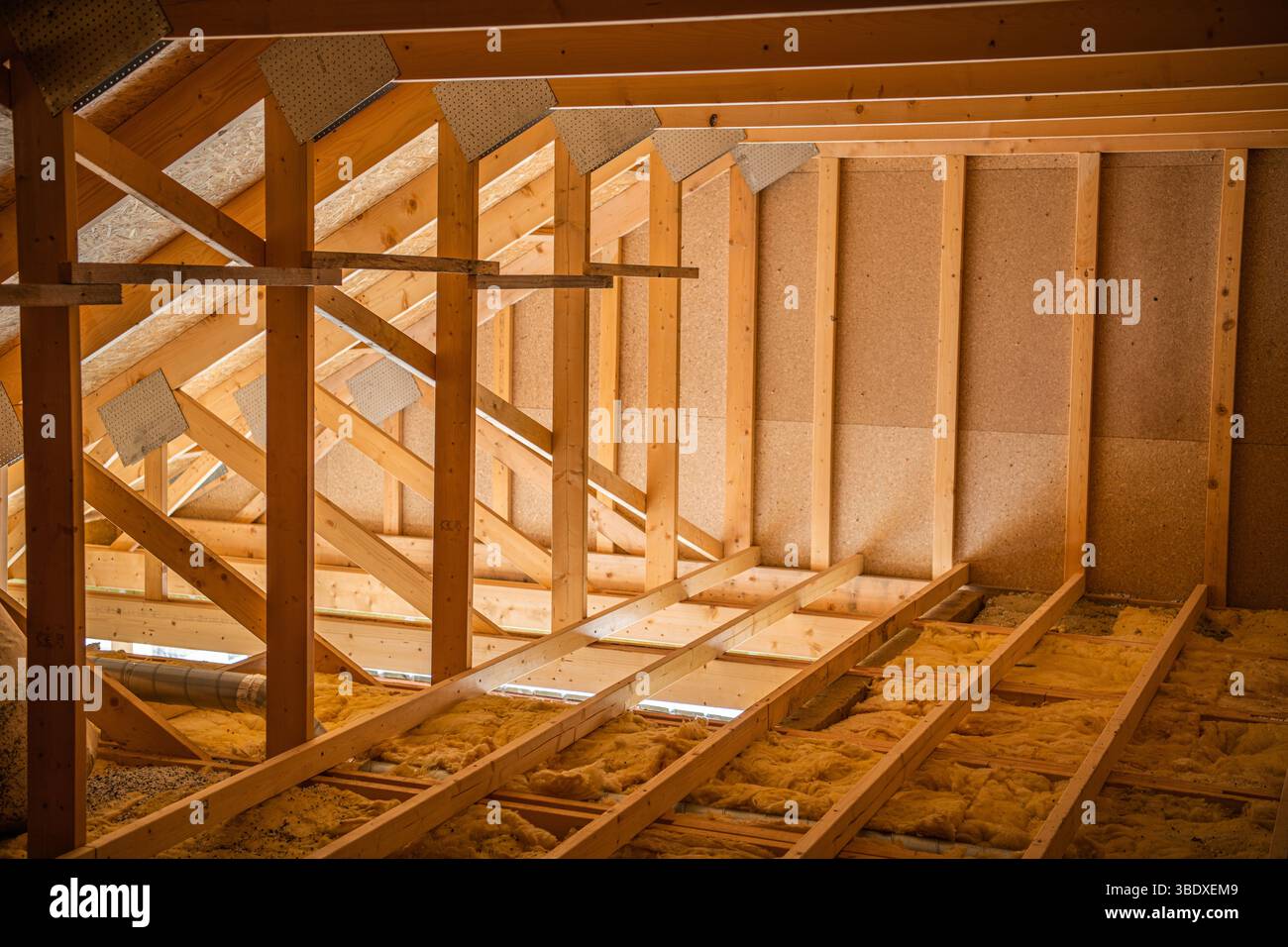 Des poutres en bois encadrent un espace mansardé pendant la construction, avec une lumière vive filtrant à travers les ouvertures et l'isolation couvrant le sol. Banque D'Images