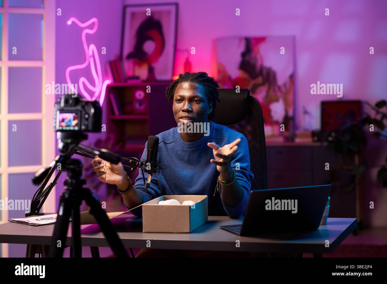 Homme afro-américain hébergeant un podcast dans un studio moderne, entouré d'équipements audio professionnels et de gadgets, engageant le public en ligne, encadré des œuvres d'art sur le mur Banque D'Images