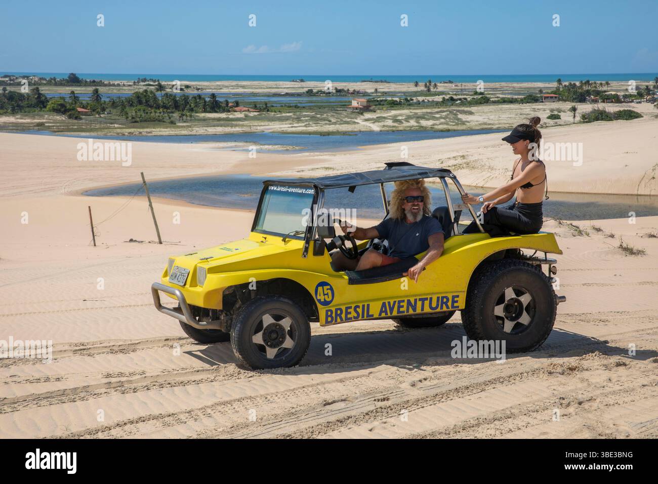 Brésil, Nordeste, Ceara, Canoa Quebrada, couple brésilien dans un buggy jaune au milieu de dunes remplies de lagons avec l'océan au loin Banque D'Images