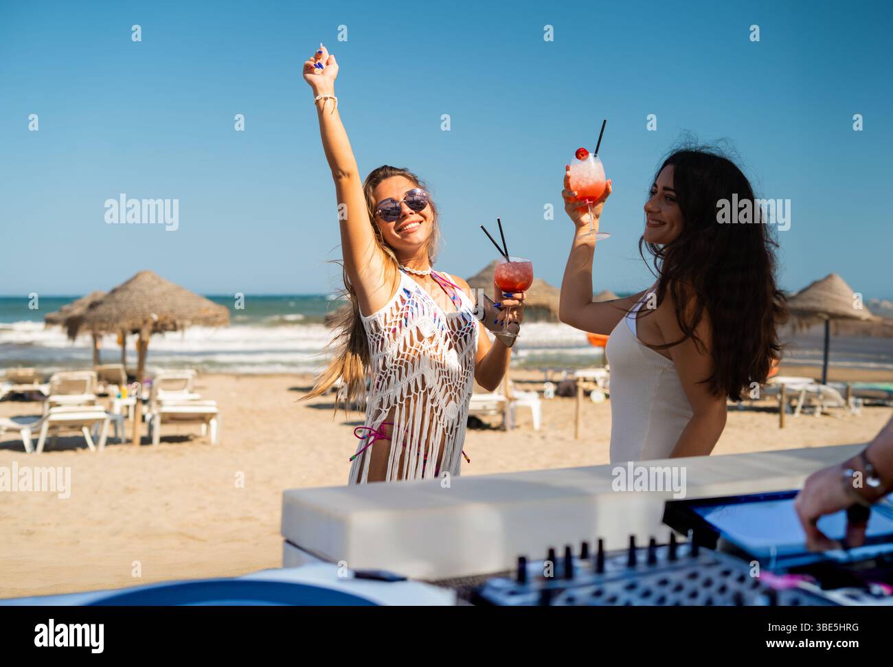 Deux amis célèbrent l'été sur une plage de sable, élevant leurs cocktails colorés dans les airs. Parapluies de paille parsèment le fond, tandis que les vagues doucement Banque D'Images