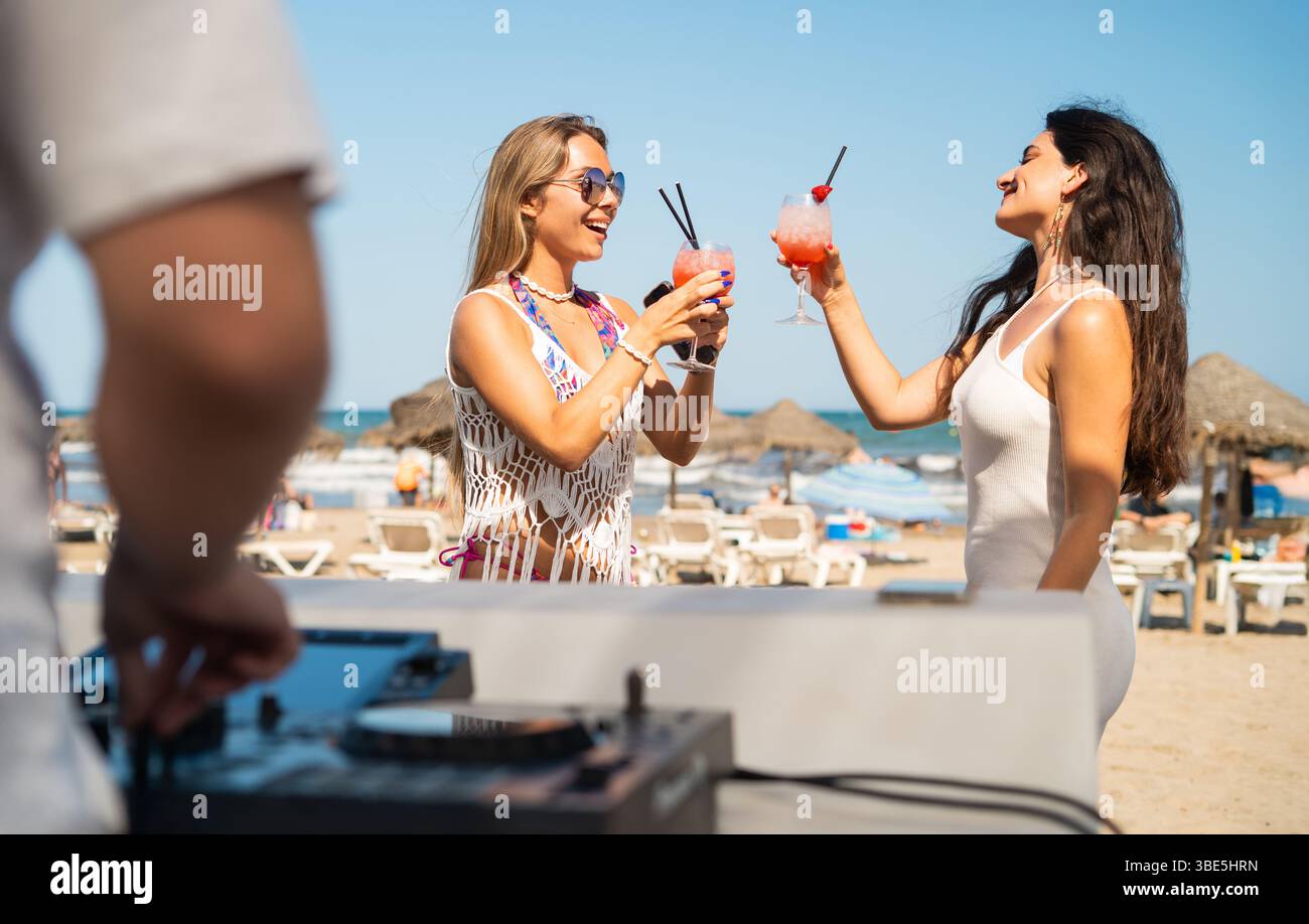 Deux femmes apprécient des cocktails colorés lors d'une fête animée sur la plage. Le cadre ensoleillé, la musique de DJ énergique et l'atmosphère joyeuse créent un bel été Banque D'Images