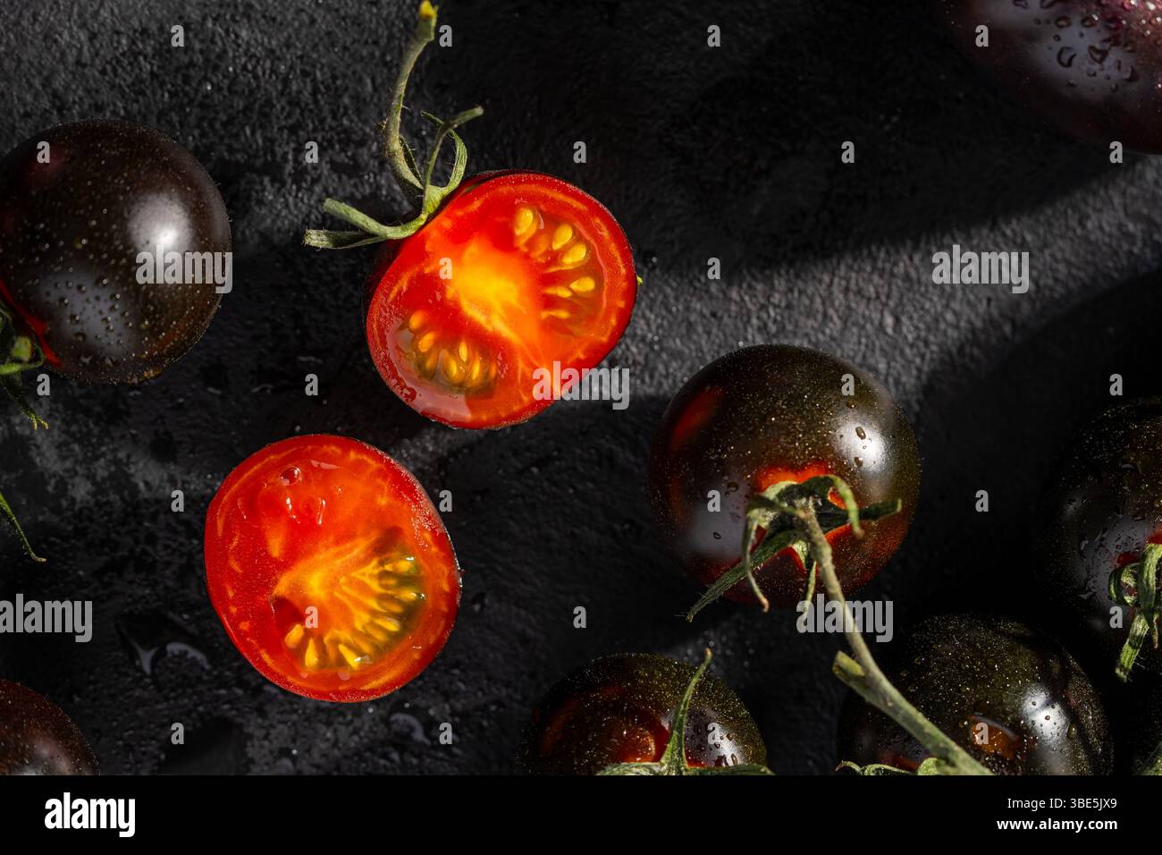 Un groupe de tomates cerises noires entières et coupées en deux, présentées sur une surface texturée sombre, mettant en valeur leur peau brillante et leur intérieur rouge vif. Banque D'Images