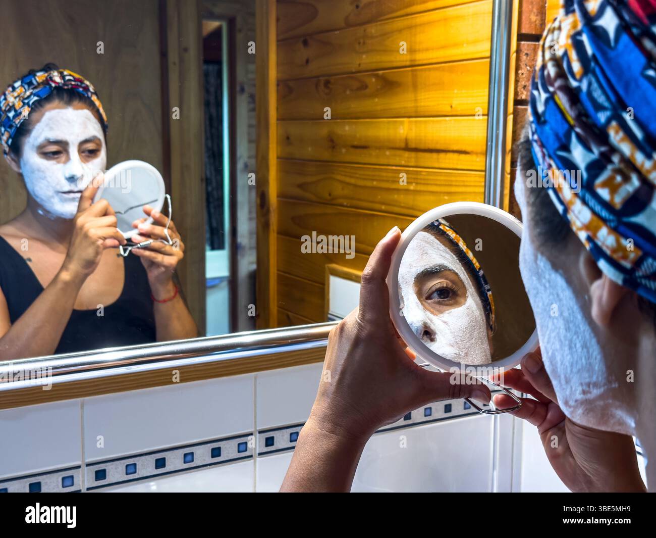 Une femme examine sa routine de soins de la peau à l'aide d'un miroir à main dans une salle de bain confortable de Victoria, en Australie. Elle porte un masque facial blanc et une cicatrice de tête colorée Banque D'Images