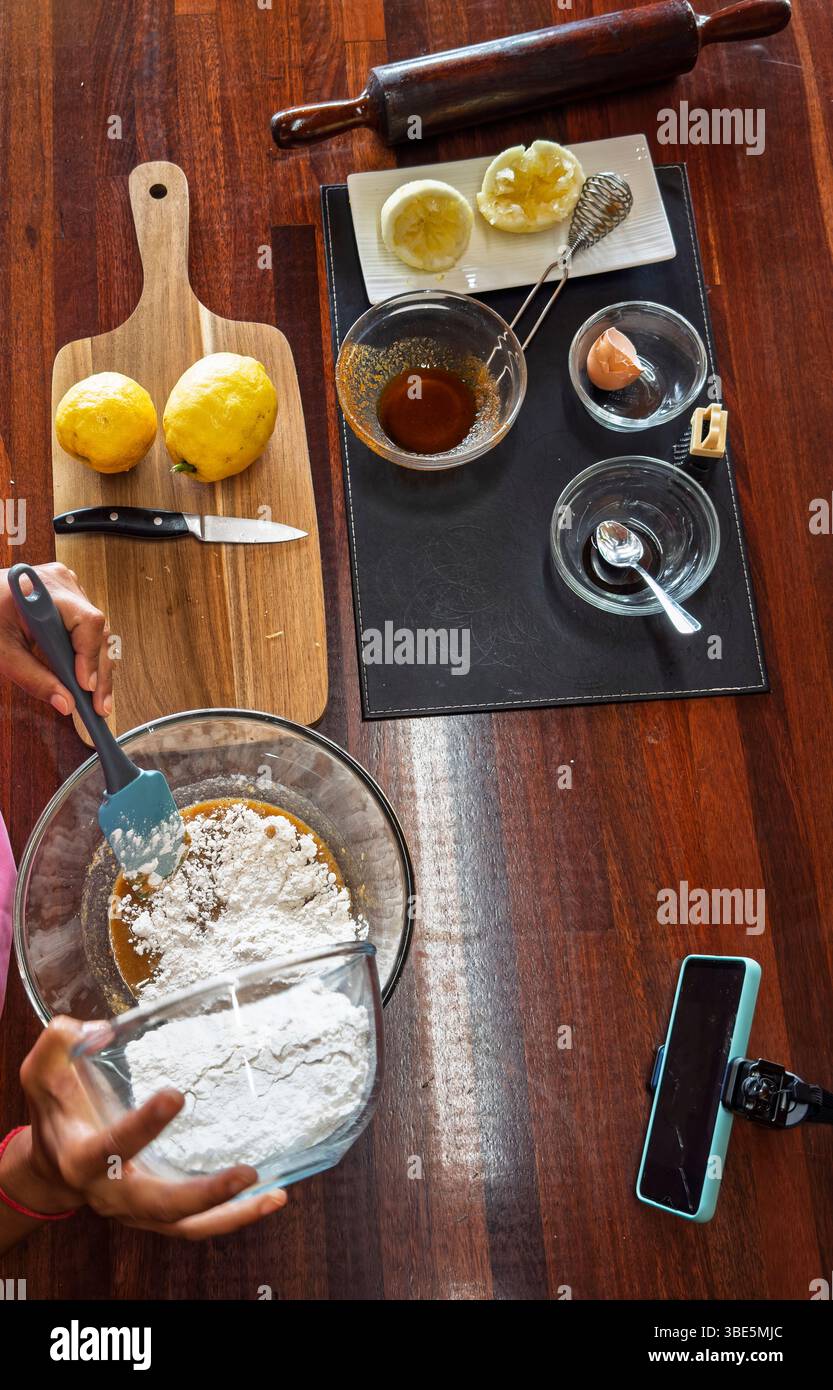 Une femme prépare la pâte à biscuits pour la cuisson dans un récipient en verre sur une table en bois, entourée de citrons, de farine, d'un rouleau à pâtisserie, d'un couteau, des œufs, et un smar Banque D'Images