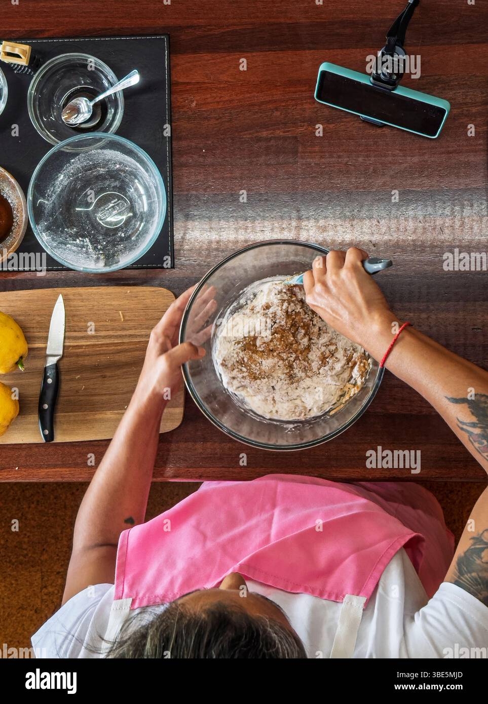 Une vue aérienne d'une femme mélangeant la pâte à biscuits dans un bol en verre sur un comptoir en bois. Divers ustensiles de cuisine, ingrédients, et une planche à découper Wit Banque D'Images
