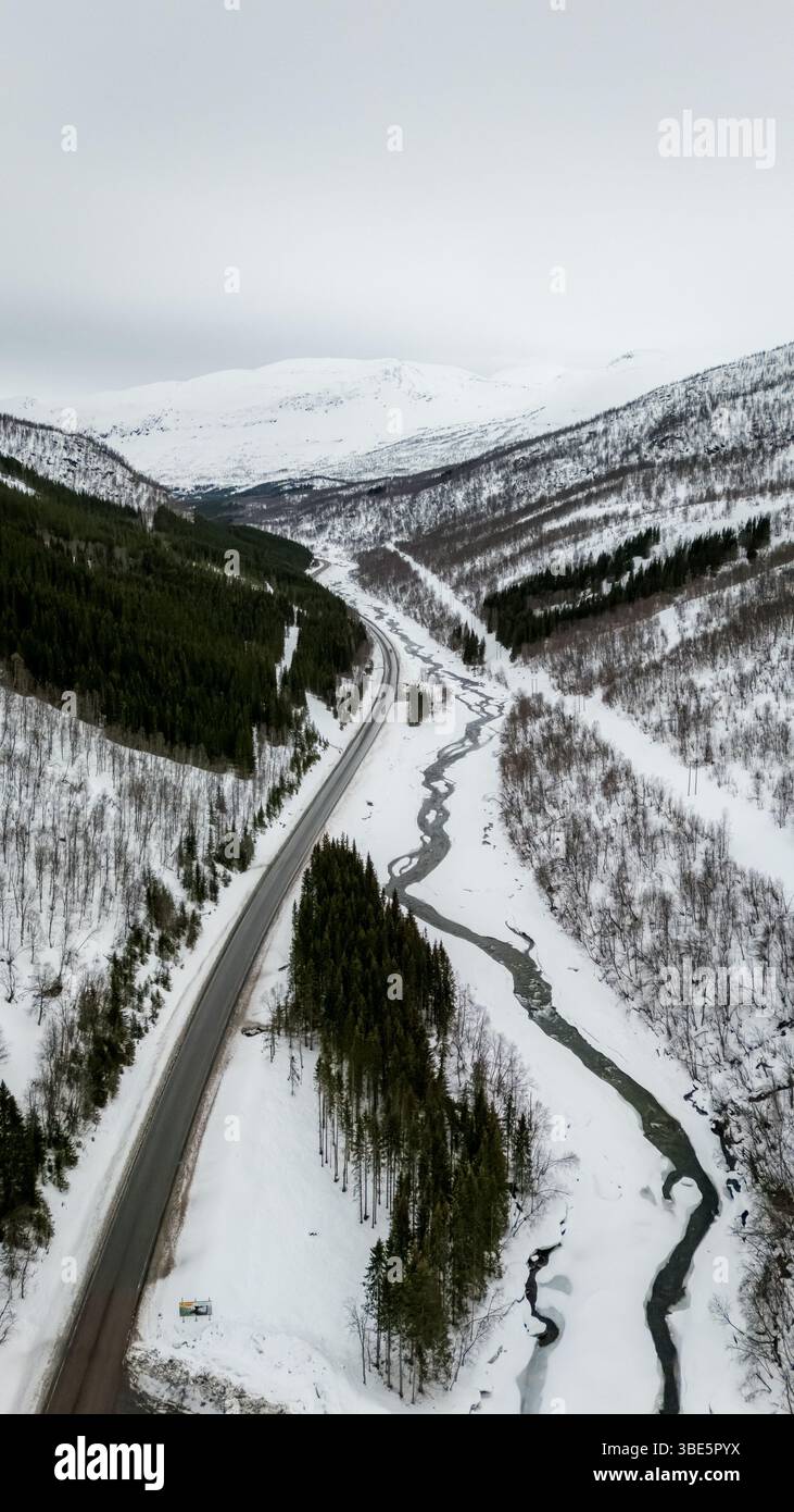 Vue aérienne d'un paysage nordique avec une route traversant des montagnes et des forêts enneigées, mettant en valeur la beauté sereine d'un pays des merveilles hivernales Banque D'Images