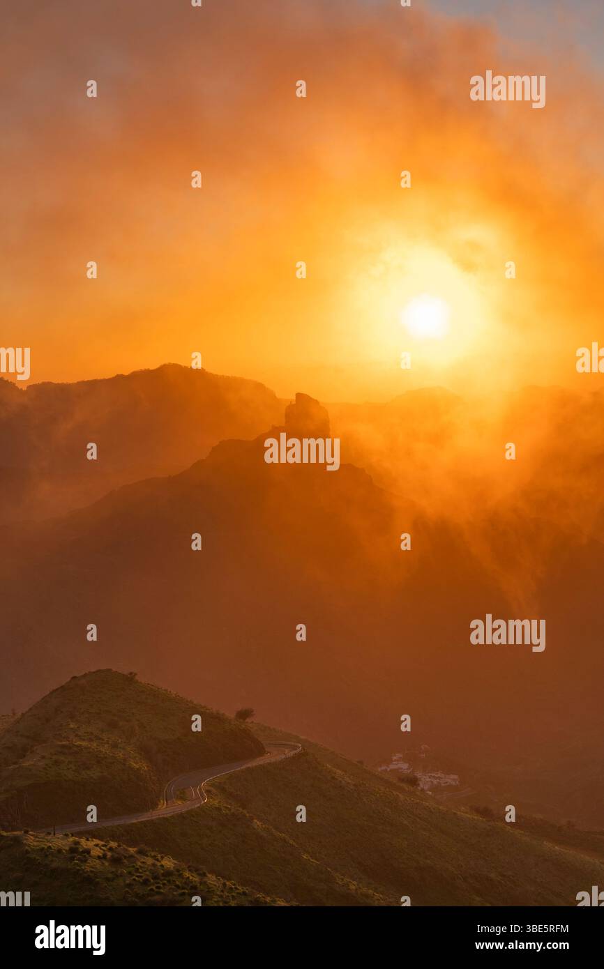 Une image frappante mettant en vedette Roque Nublo enveloppée dans une brume dorée au coucher du soleil, avec une route sinueuse en pente douce à travers le terrain accidenté. Banque D'Images