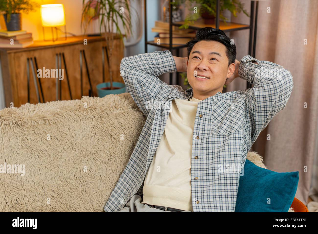 Heureux homme chinois d'âge moyen insouciant assis confortablement avec la main derrière la tête sur le canapé dans le salon à la maison. Souriant mec coréen se relaxant sur le canapé pensant regarder autour dans la chambre de l'appartement. Banque D'Images