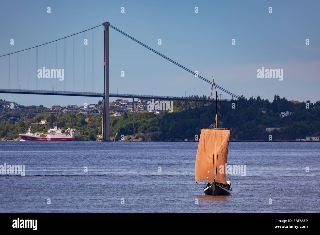 Voilier traditionnel en bois avec voile rouge passant sous un pont suspendu moderne en Norvège côtière par une journée d'été claire. Banque D'Images Voilier traditionnel en bois avec voile rouge passant sous un pont suspendu moderne en Norvège côtière par une journée d'été claire. Banque D'Images
