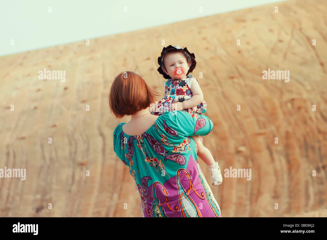 Mère tenant sa petite fille dans les bras, debout dehors contre une colline sablonneuse. Amour de famille, liens d'été et concept de parentalité dans la lumière naturelle Banque D'Images