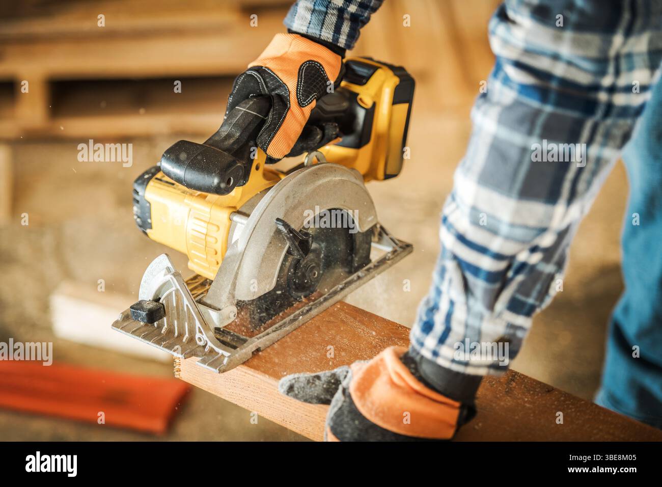 Un menuisier se concentre sur la découpe d’une planche de bois avec une scie circulaire dans un atelier bien éclairé. Les particules de poussière sont visibles dans l'air pendant sa concentration. Banque D'Images