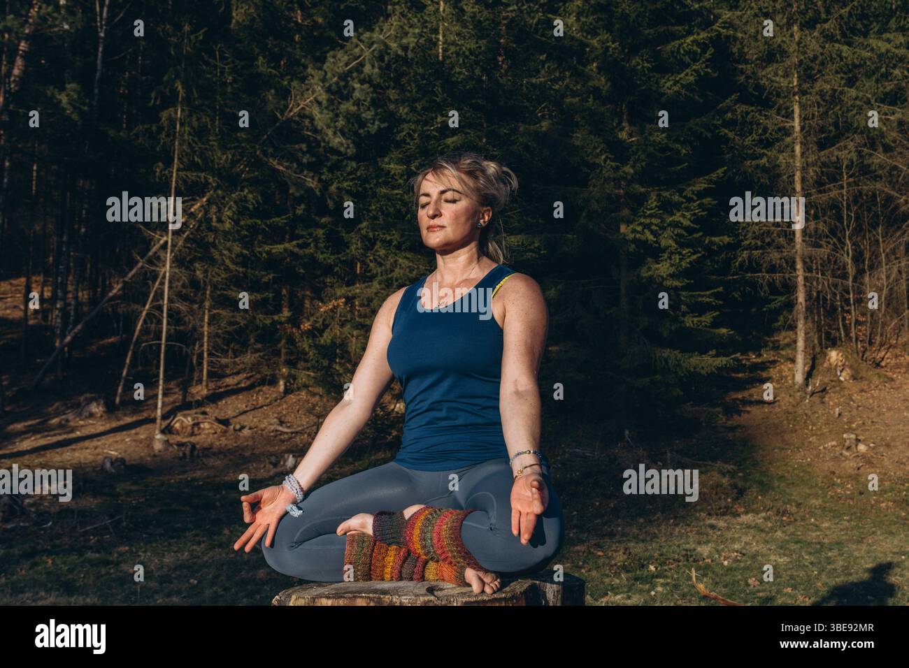 Une femme pratiquant le yoga effectue une séance assise en position lotus sur une souche d'arbre, méditant seule dans des vêtements décontractés lors d'une journée d'automne ensoleillée dans le parc. Bonjour Banque D'Images