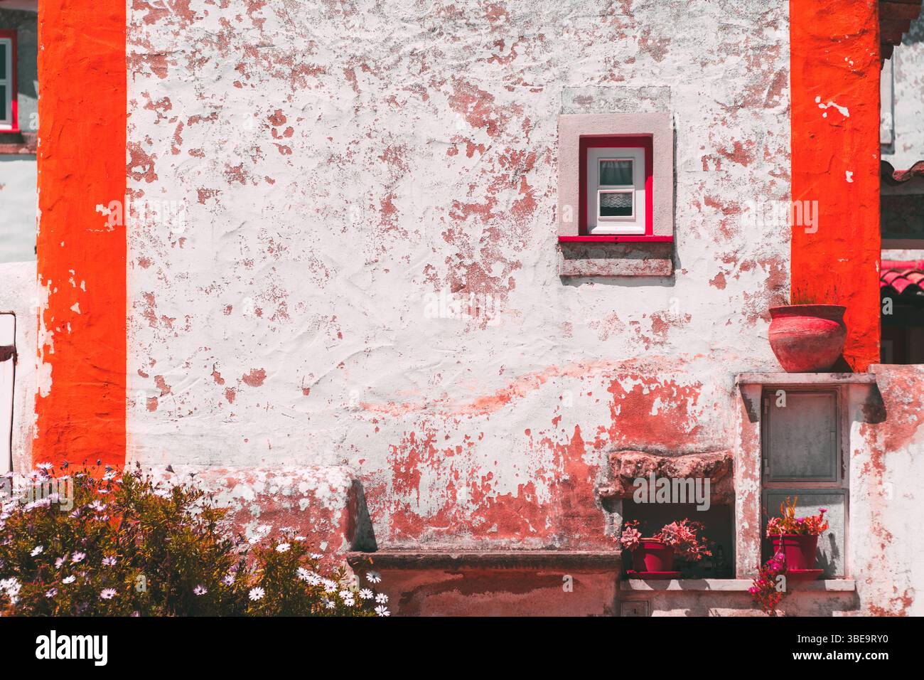 Charmante façade rustique d'une ancienne maison méditerranéenne avec des murs texturés blancs et rouges altérés par les intempéries, de petites fenêtres, des pots en argile et des fleurs en fleurs sous Banque D'Images