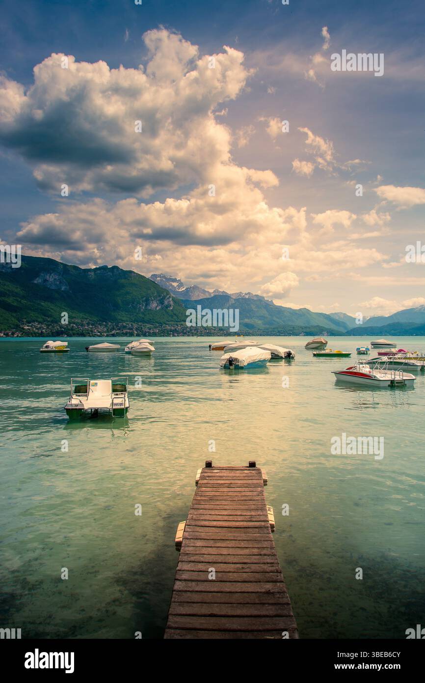 Un ponton en bois s’étend sur les eaux tranquilles du lac d’Annecy, entouré de montagnes et parsemé de bateaux colorés. Haute Savoie. France Banque D'Images