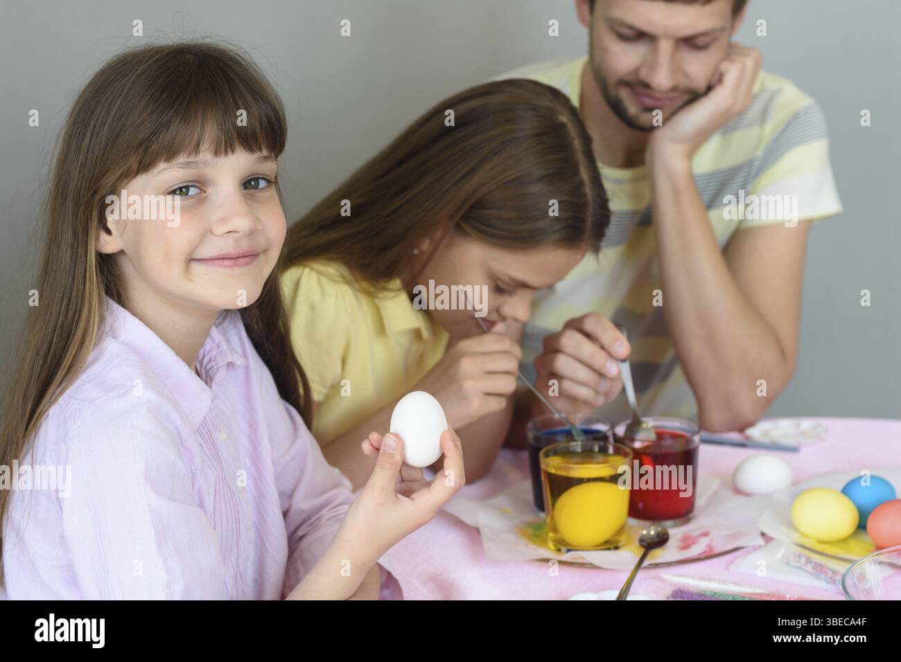 Une fille avec un oeuf dans ses mains regardait dans le cadre, à l'arrière-plan la famille peint des oeufs dans des verres avec des colorants Banque D'Images