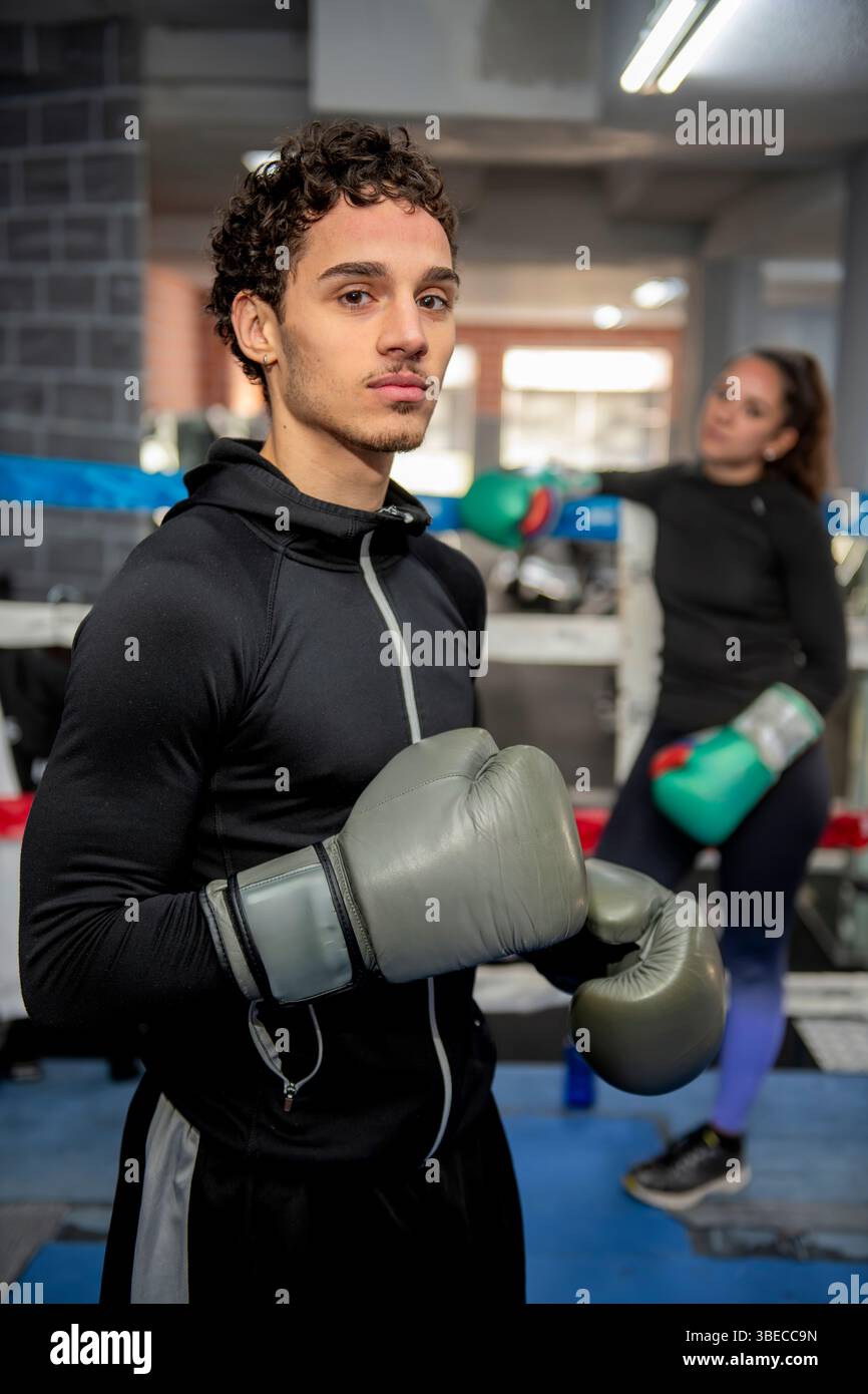 Jeune boxeur masculin hispanique portant des gants de boxe posant dans un anneau de gymnastique avec boxer féminin en arrière-plan Banque D'Images