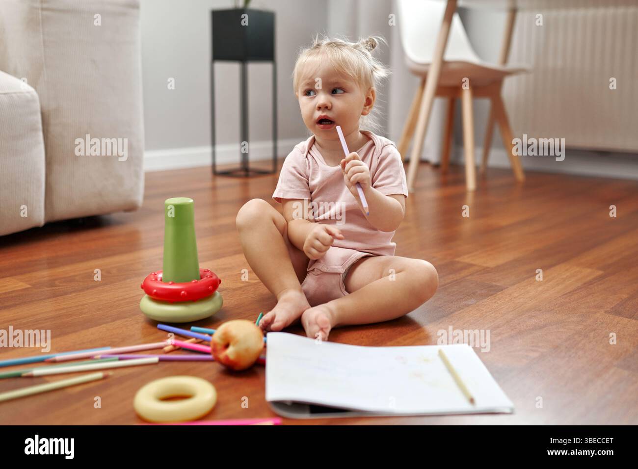 Un tout-petit joyeux est assis sur le sol mou, entouré de jouets colorés, une pomme rouge brillante et des matériaux de dessin, profitant d'un p délicieux et créatif Banque D'Images