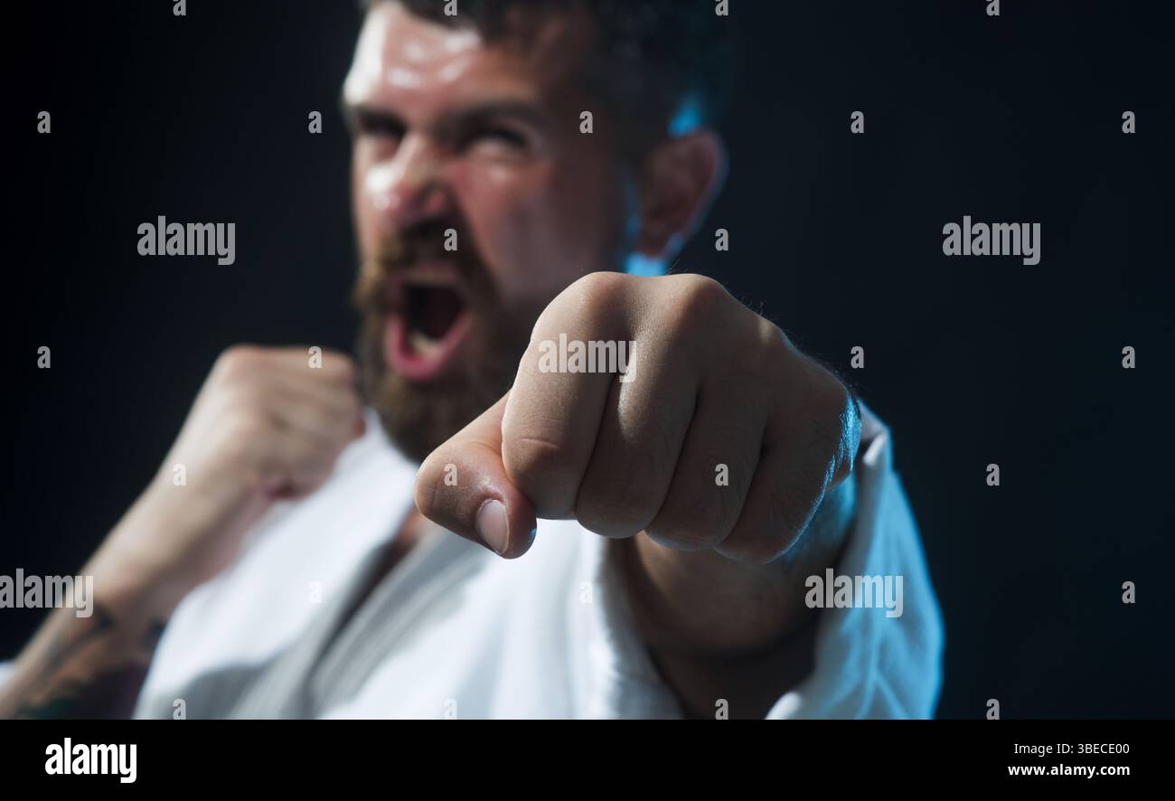 Karaté homme en posture de combat. Combattant de karaté hurlant pratiquant des coups de poing dans la salle de gym, focus sélectif sur le poing. Homme barbu en colère en kimono avec clen de main Banque D'Images