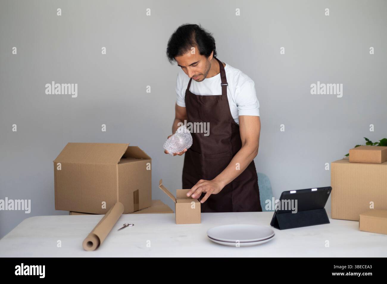 Homme enveloppant un article dans du papier bulle et le plaçant dans une petite boîte, préparant des paquets sur une table avec des boîtes et des assiettes Banque D'Images