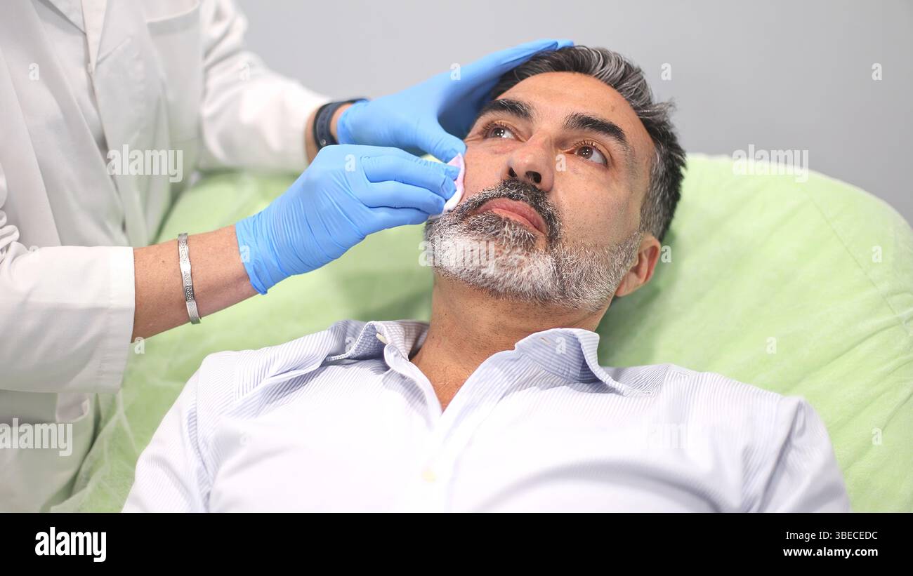 Médecin nettoyant le visage d'un patient masculin avec un chiffon après une procédure cosmétique dans une clinique de beauté, patient allongé et levant les yeux. Banque D'Images
