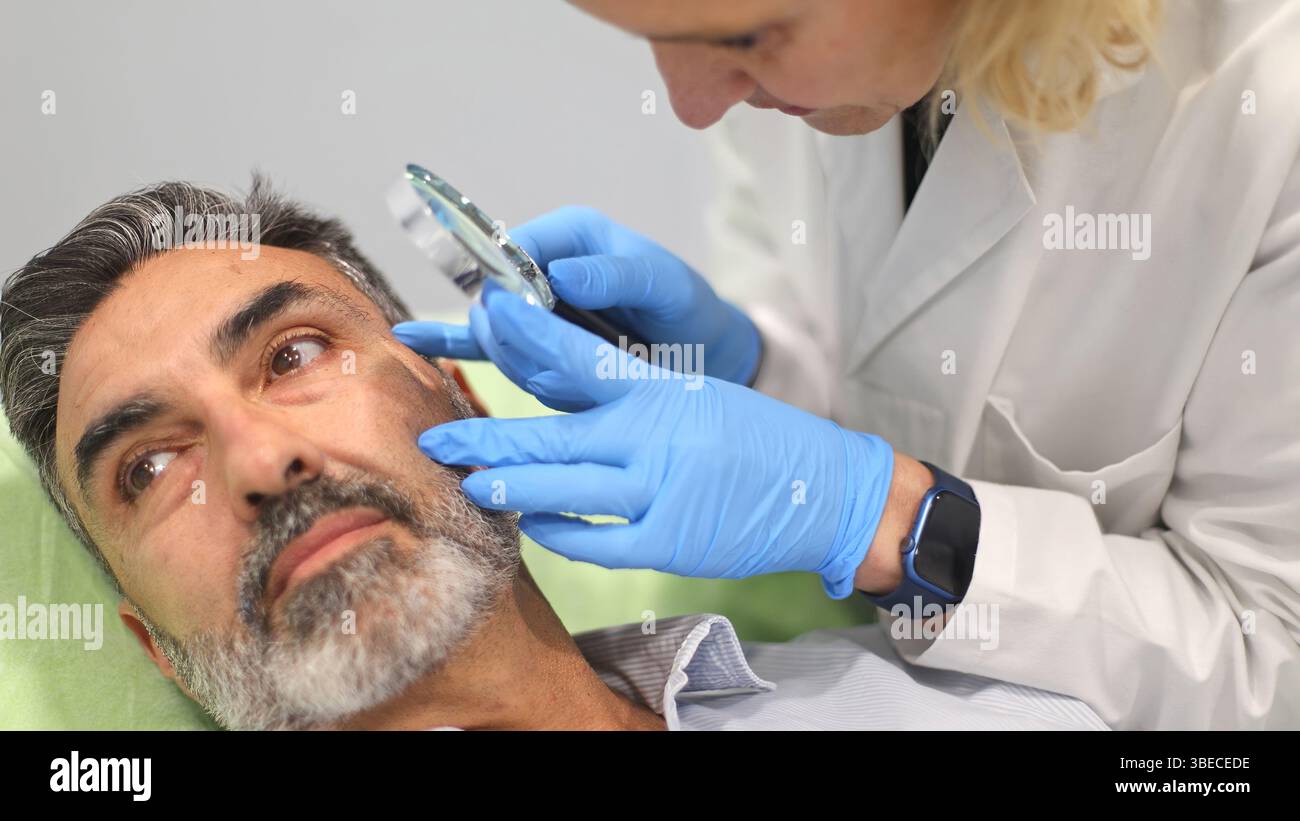 Médecin inspectant le visage d'un patient masculin avec une loupe après une procédure cosmétique dans une clinique de beauté, patient allongé. Banque D'Images