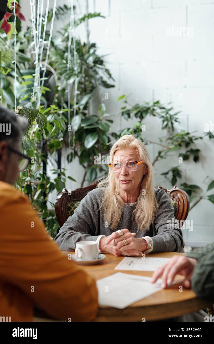 Femme âgée avec des cheveux blonds et des lunettes conversant avec deux hommes dans un cadre intérieur confortable entouré de plantes vertes illuminant le fond. Femme souriante et engagée dans la discussion Banque D'Images