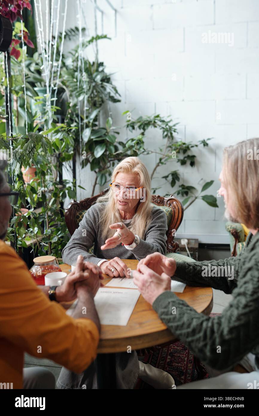 Trois personnes ont participé à une discussion animée dans un café intérieur confortable avec des plantes et un éclairage chaleureux, une personne âgée portant des lunettes axées sur la communication de leurs idées Banque D'Images