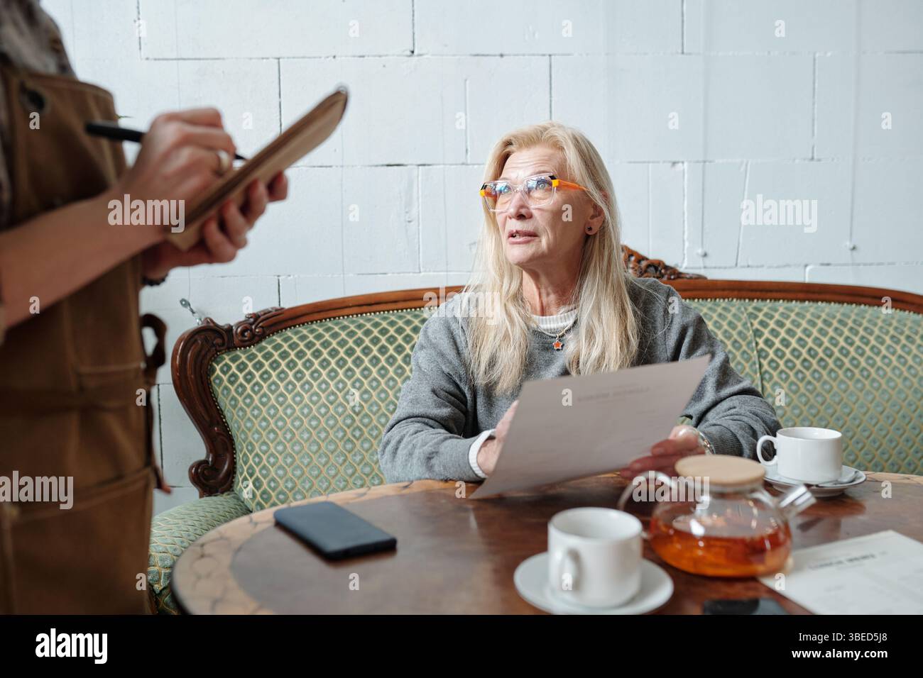 Femme âgée tenant le document pendant la réunion dans un café confortable, s'engageant dans la conversation avec une autre personne prenant des notes. Table en bois chaude avec tasses à thé et théière entre eux Banque D'Images