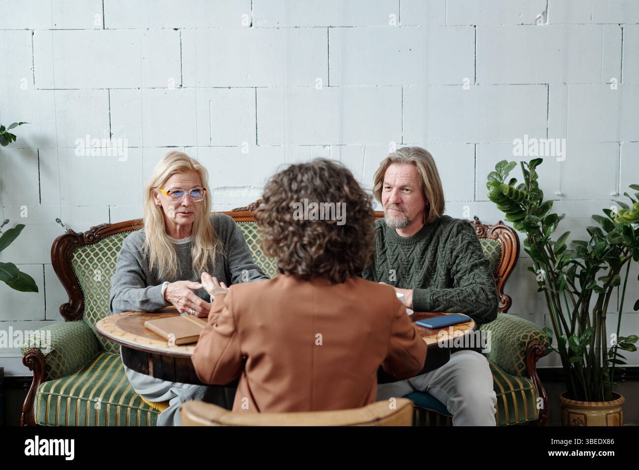 Couple âgé s'engageant dans la conversation avec une autre personne à la table basse ronde, entouré de plantes vertes et de meubles élégants Banque D'Images