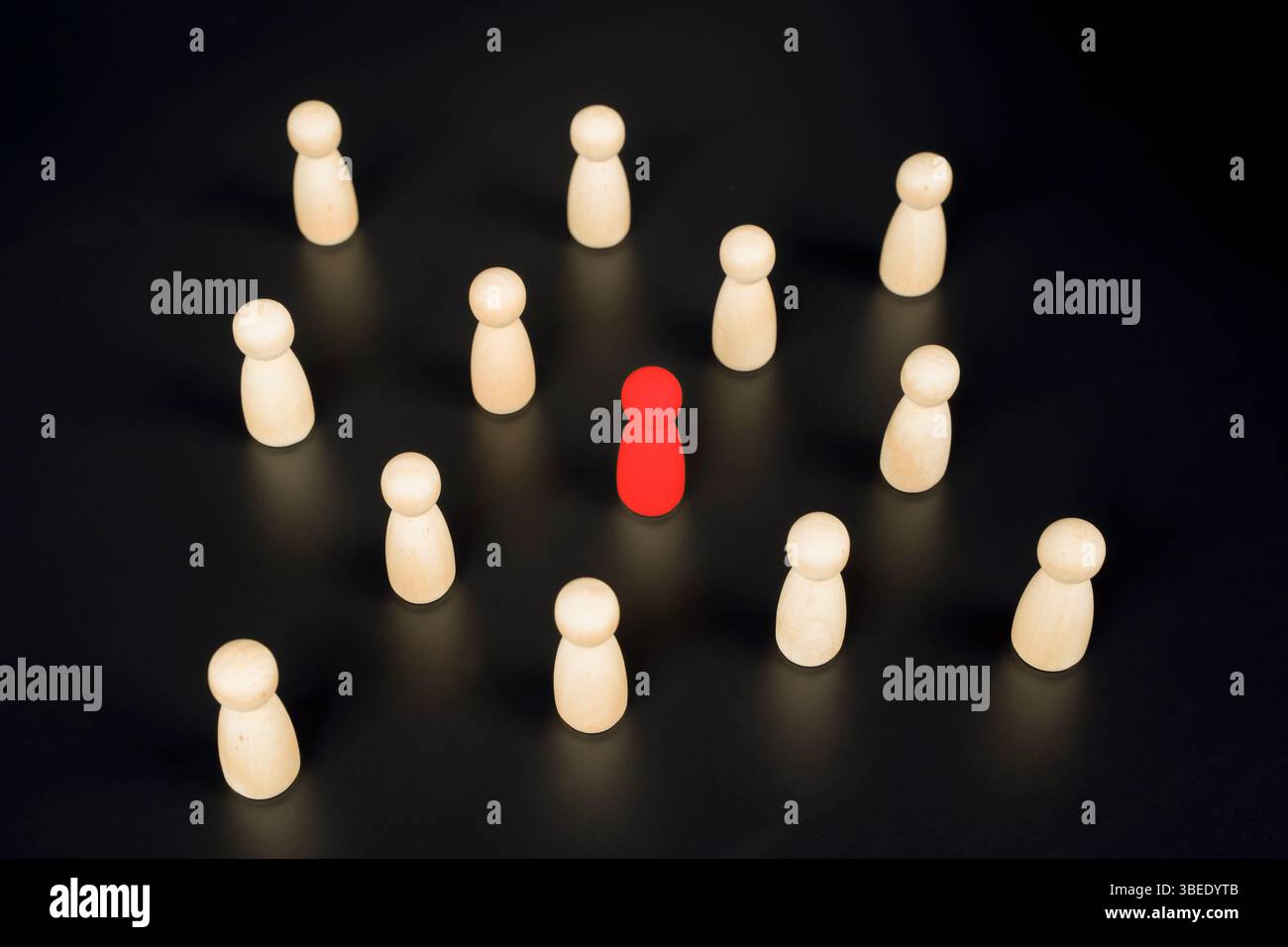 Une pièce de jeu rouge est isolée au centre, entourée de figures en bois identiques sur fond noir, symbolisant la solitude, l'exclusion, le social Banque D'Images