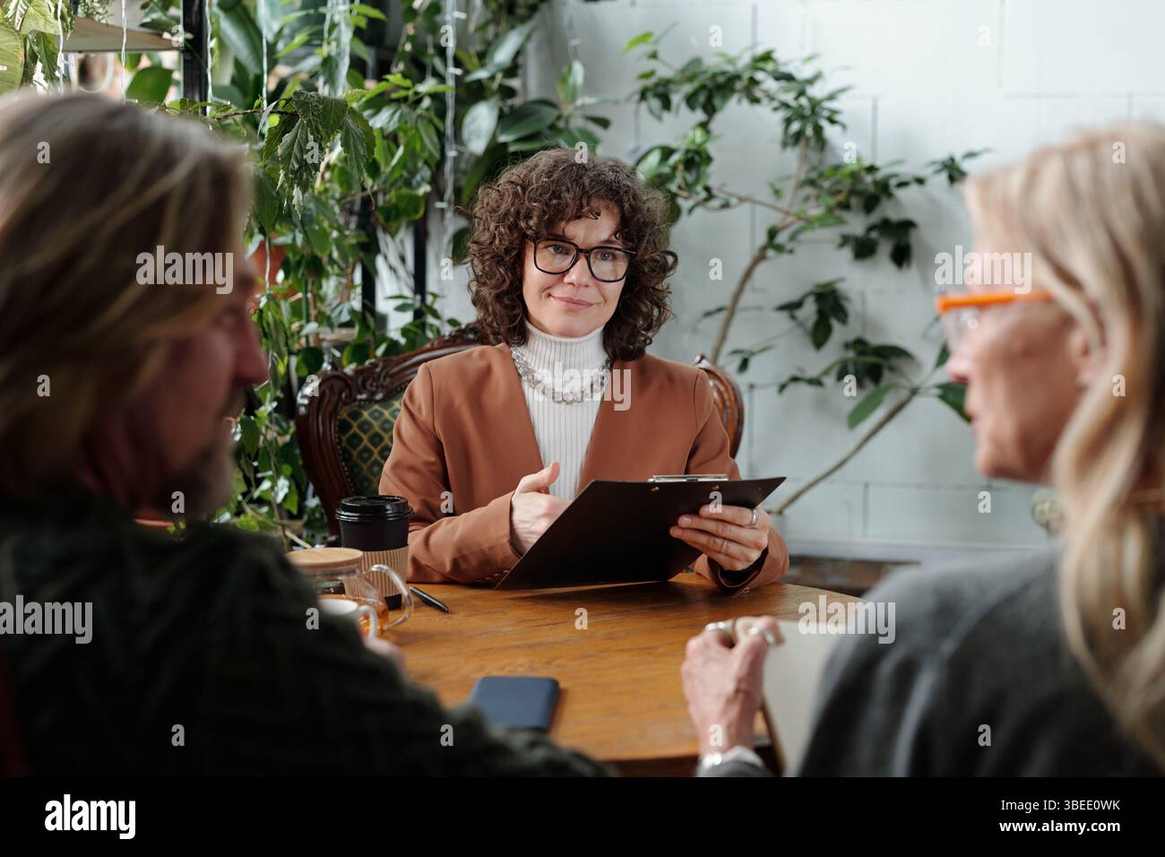 Femme d'âge moyen avec des cheveux bouclés tenant la tablette, s'engager avec des collègues pendant la réunion informelle. Scène intérieure avec des plantes en arrière-plan Banque D'Images