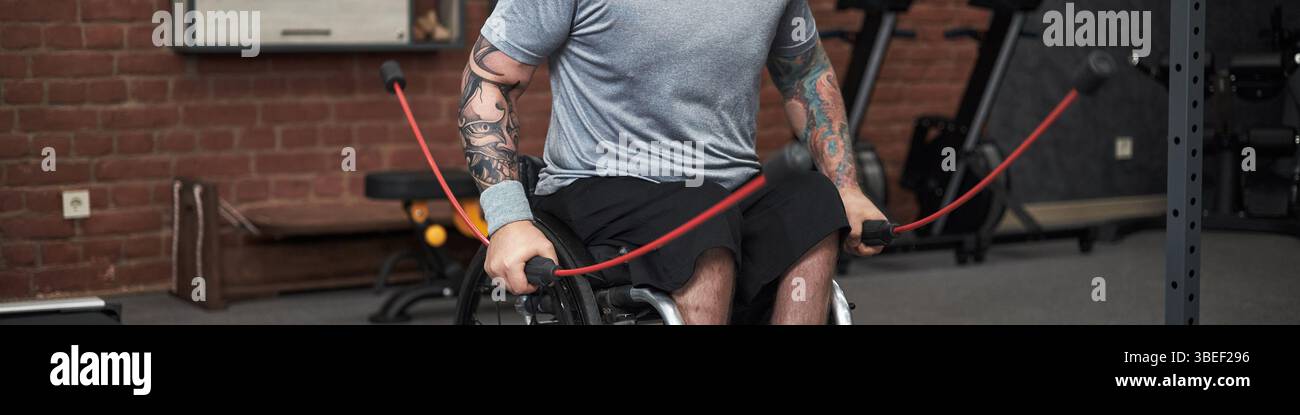 En-tête avec l'homme dans la salle de gym engagé dans l'entraînement de force avec des bandes de résistance, en mettant l'accent sur l'entraînement du haut du corps. Tatouages détaillés visibles sur les bras, équipement de gymnastique Banque D'Images