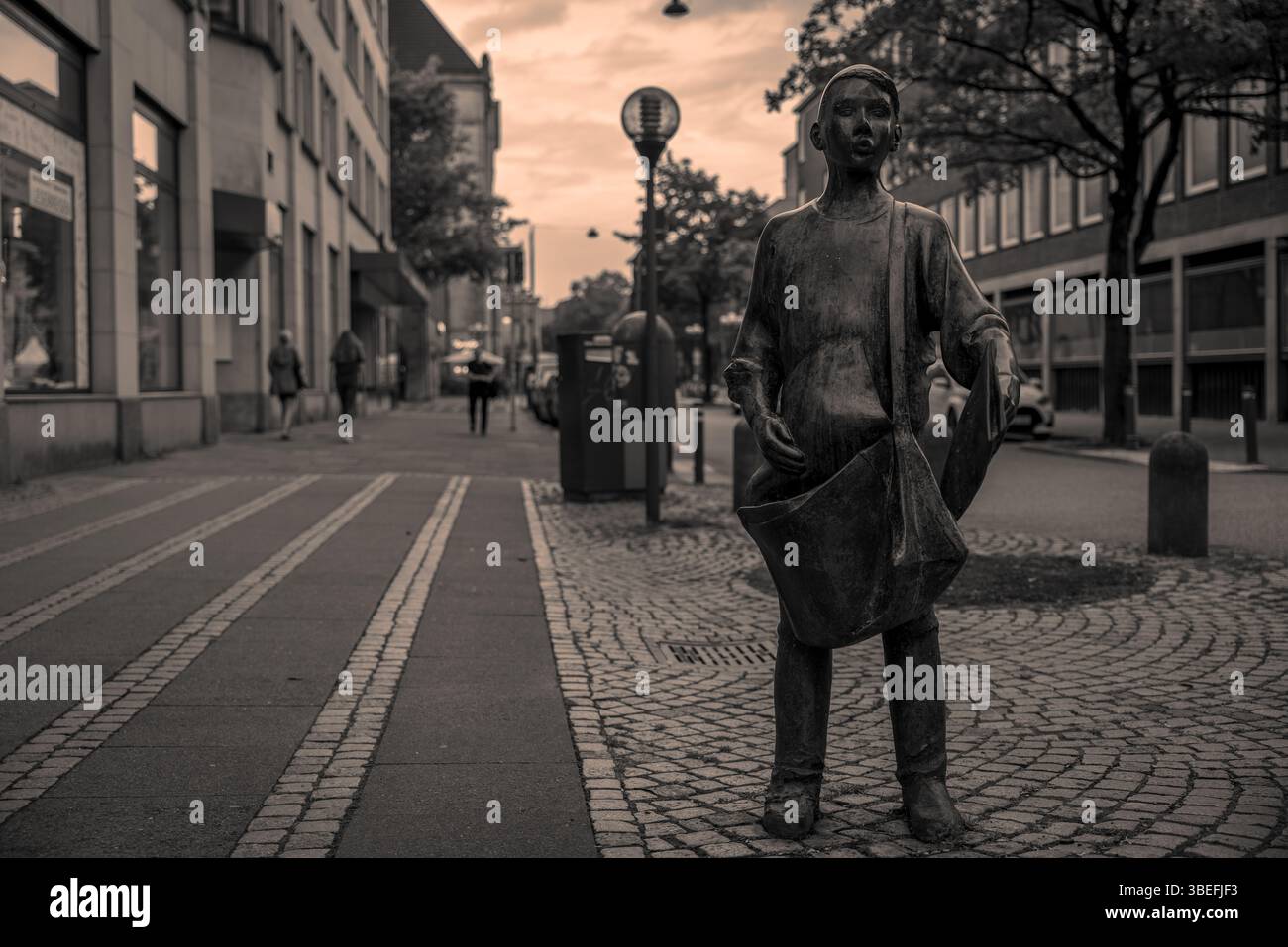 Vue monochrome de la statue de Newsboy en bronze sur Holstenstraße, Kiel, Allemagne, avec des piétons et des boutiques en arrière-plan doux. Éditorial uniquement. Banque D'Images