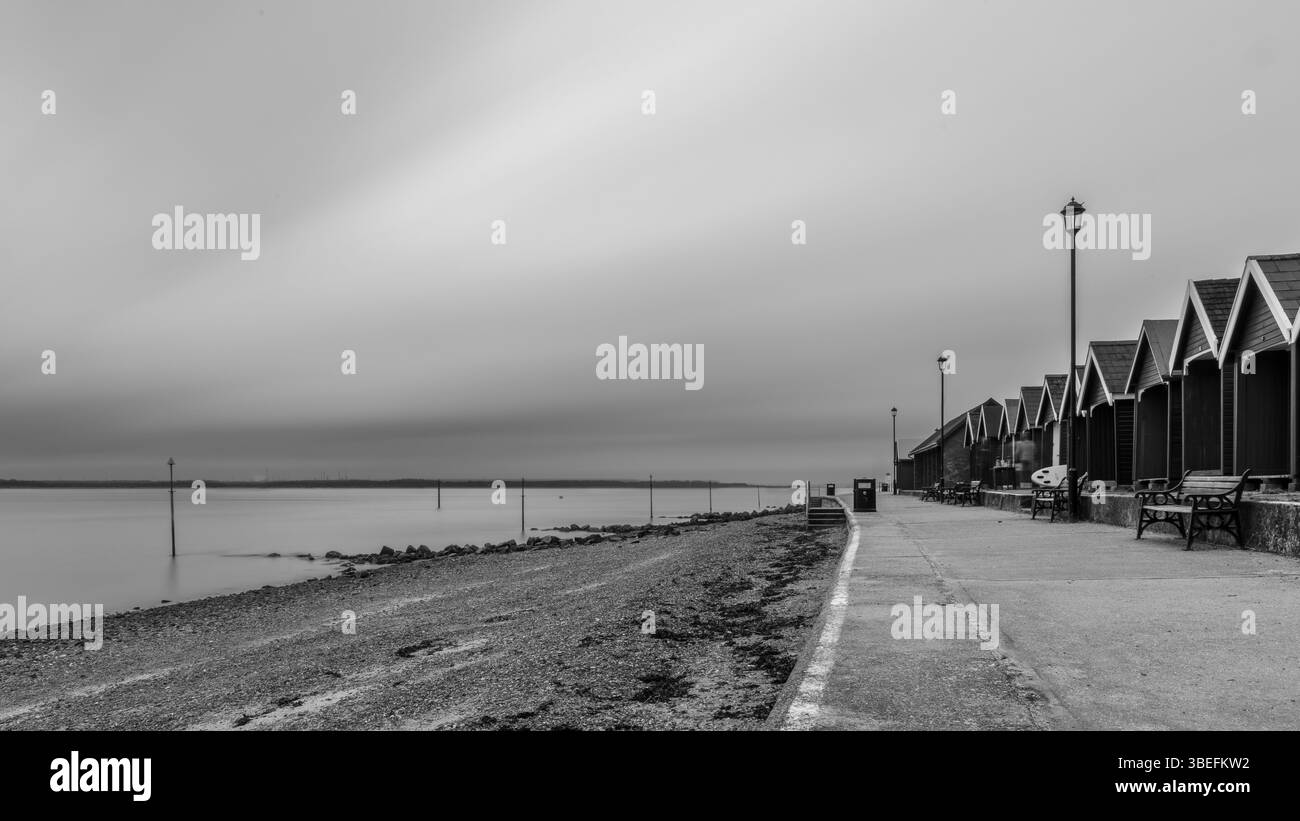 Image en noir et blanc de cabanes de plage le long du front de mer de Gurnard sur l'île de Wight, avec une mer lisse et des bancs vides par une journée brumeuse. Banque D'Images Image en noir et blanc de cabanes de plage le long du front de mer de Gurnard sur l'île de Wight, avec une mer lisse et des bancs vides par une journée brumeuse. Banque D'Images