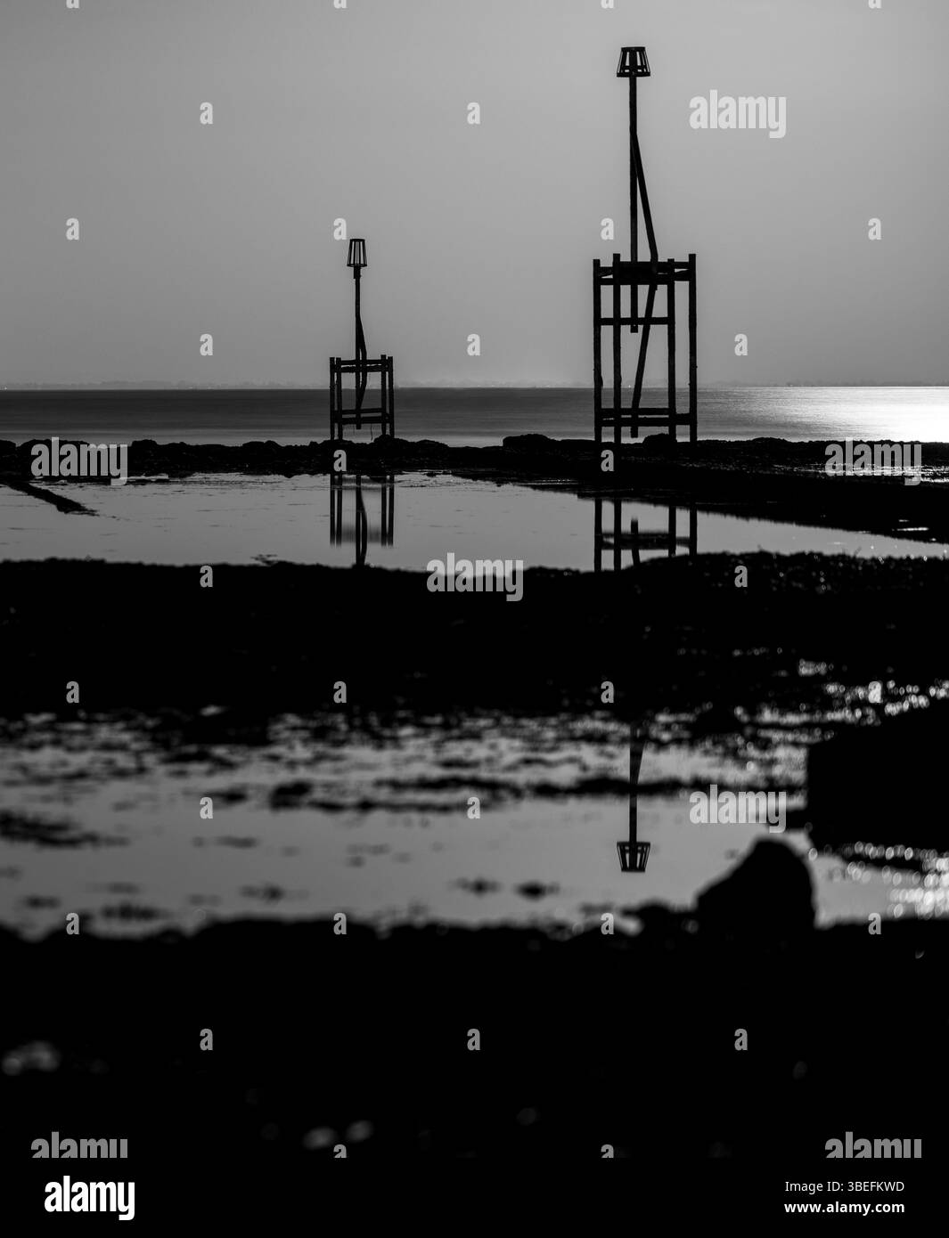 Des marqueurs de marée silhouettés se dressent à marée basse sur l'île de Wight, avec des reflets visibles en eau calme et un horizon minimaliste. Banque D'Images Des marqueurs de marée silhouettés se dressent à marée basse sur l'île de Wight, avec des reflets visibles en eau calme et un horizon minimaliste. Banque D'Images