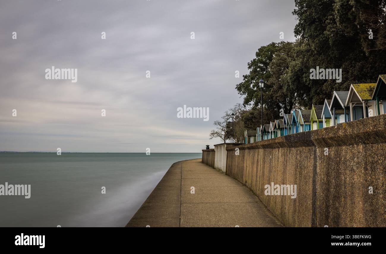 Une promenade légèrement incurvée avec des cabanes de plage colorées le long de la digue sur l'île de Wight, capturée avec une longue exposition pour lisser la mer. Banque D'Images Une promenade légèrement incurvée avec des cabanes de plage colorées le long de la digue sur l'île de Wight, capturée avec une longue exposition pour lisser la mer. Banque D'Images