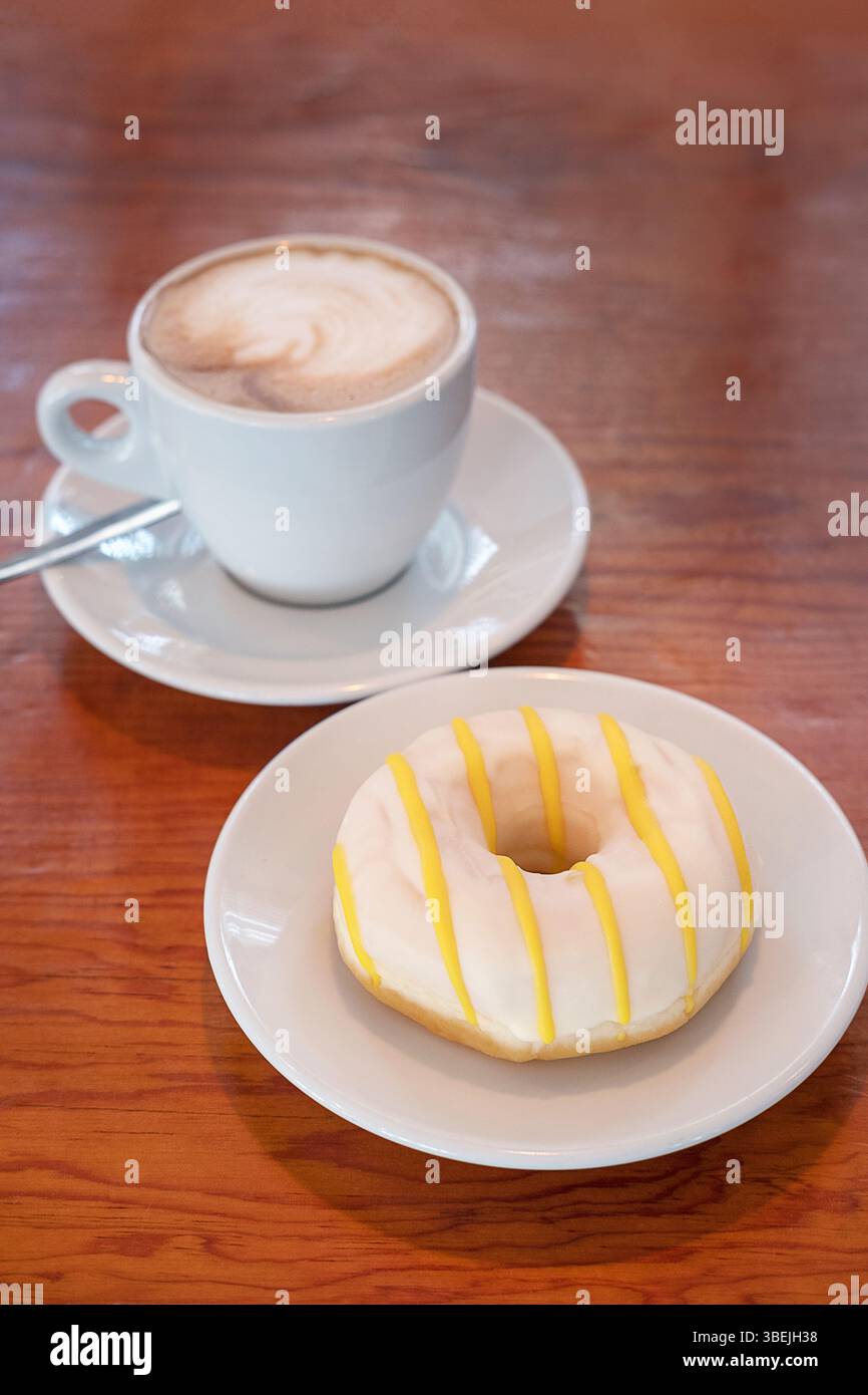 Tasse de cappuccino en céramique avec latte art servie sur une soucoupe avec une cuillère accompagnée d'un beignet blanc givré frissé avec un glaçage jaune vif Banque D'Images