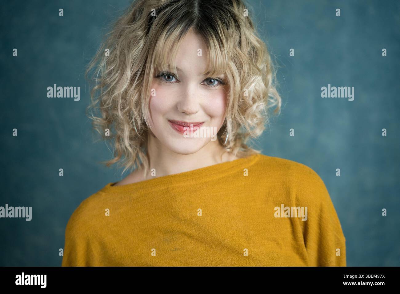 Une jeune femme souriante aux cheveux blonds bouclés et à la frange porte un pull jaune moutarde. Elle se tient debout sur un fond bleu, exsudant chaleur et con Banque D'Images