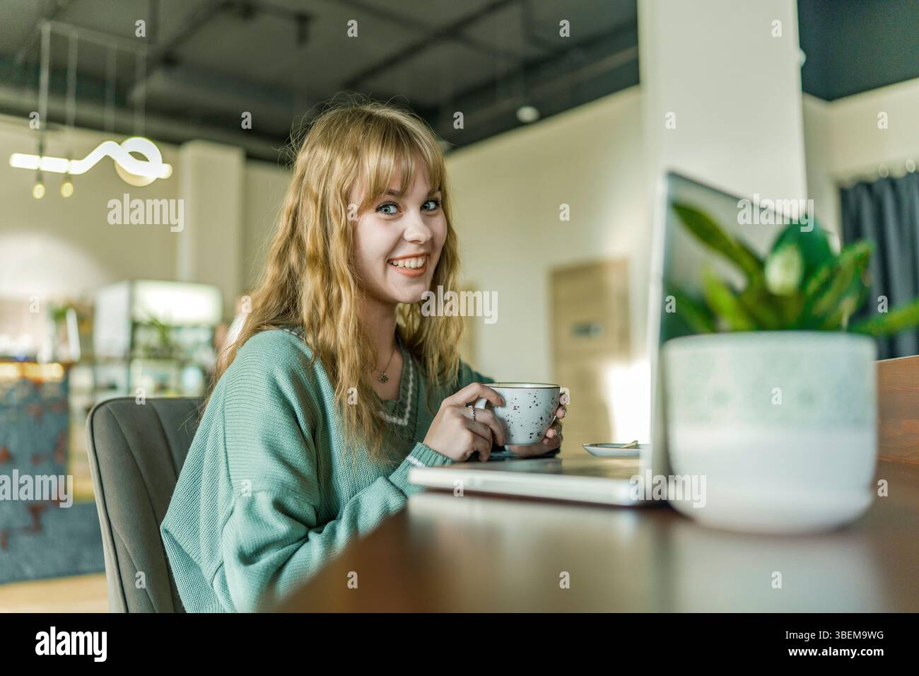 Une jeune fille blonde joyeuse appréciant son café dans un café confortable. Elle porte un pull vert clair, souriant chaleureusement tout en tenant une tasse, créant un a accueillant Banque D'Images