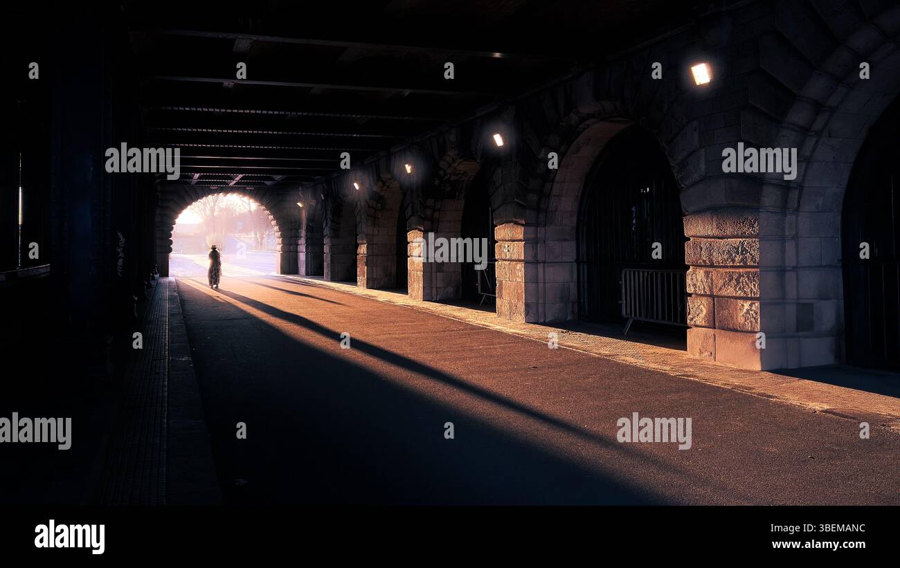 Une scène dramatique capturée dans un tunnel ou un passage souterrain, avec une série d'arches en pierre projetant de longues ombres sur le sol. Une figure solitaire marche Banque D'Images
